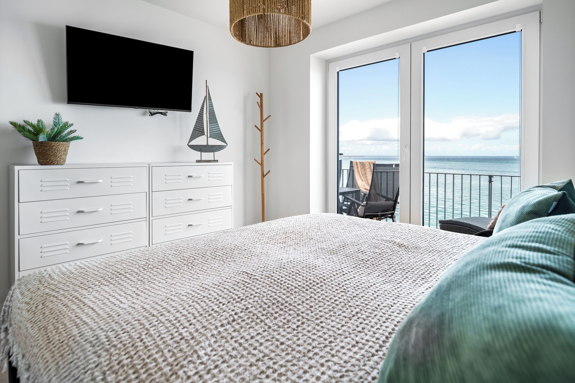 A bedroom with a large bed, a flat-screen TV, a plant, and a view of the sea through the balcony doors.