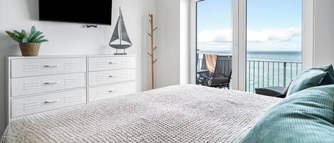 A bedroom with a large bed, a flat-screen TV, a plant, and a view of the sea through the balcony doors.