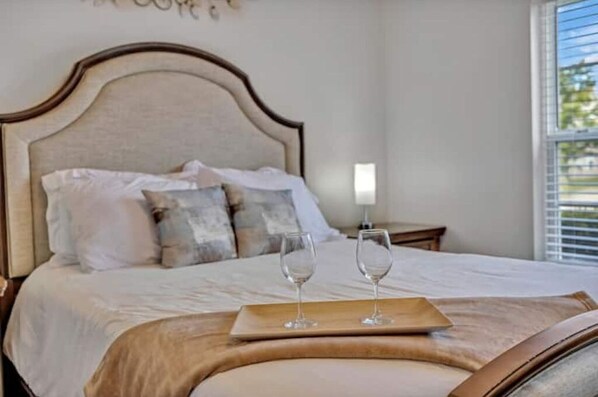 A neatly made bed with pillows and a tray with two glasses, a bedside table with a lamp, and a window with blinds.