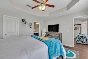 Une chambre à coucher comprenant un lit, un ventilateur de plafond, une télévision et une porte donnant sur une autre pièce.