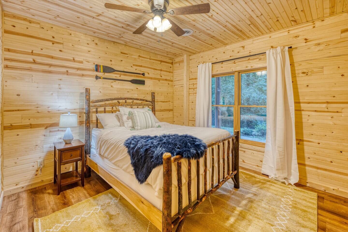 A wooden cabin bedroom with a bed, a nightstand, a lamp, a ceiling fan, and a window with curtains.