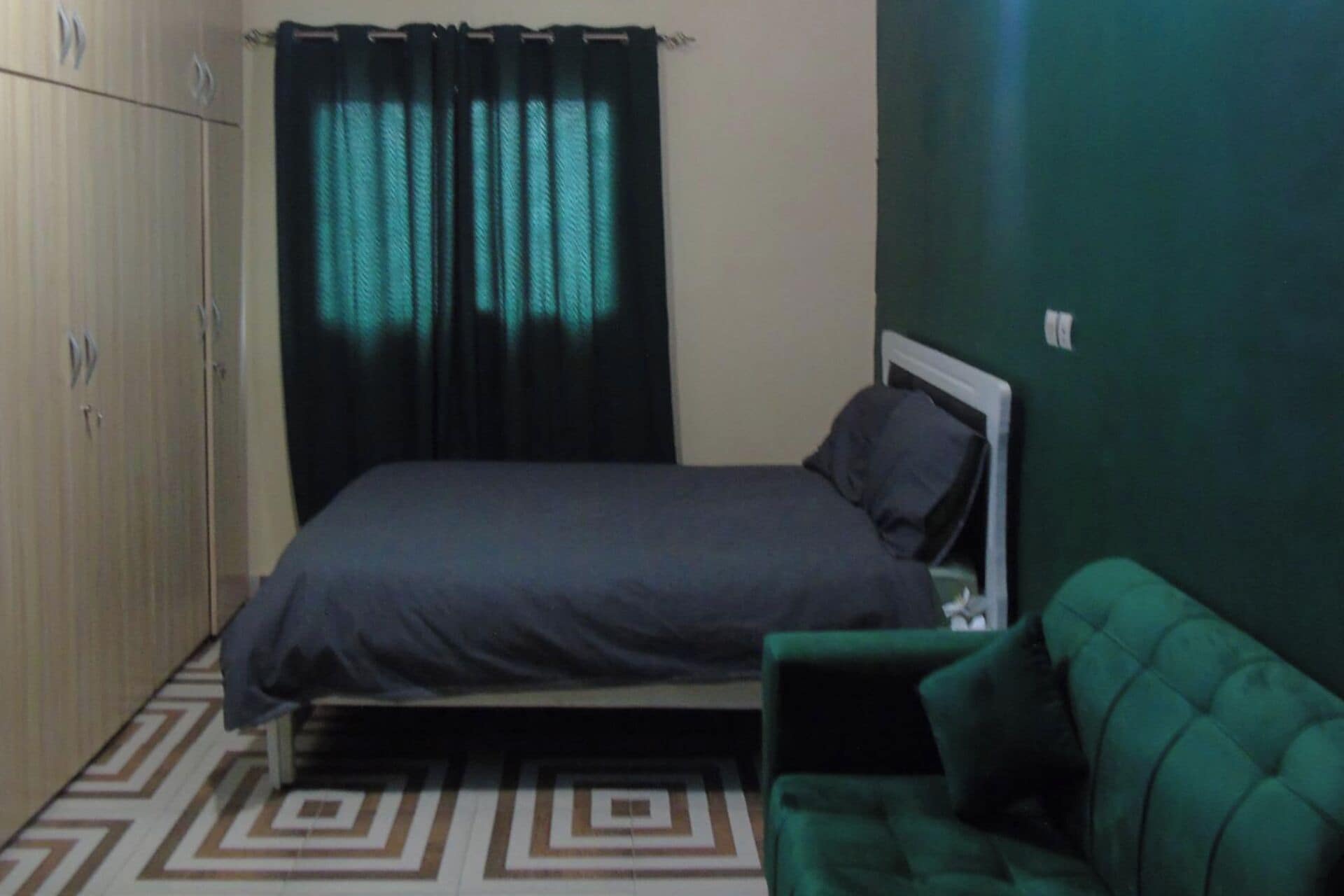 A bedroom with a bed, a green couch, a wardrobe, and a patterned floor.
