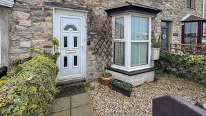 Ein Steinhaus mit weißer Tür, einem Erker und einem kleinen Gartenbereich mit Kies und Topfpflanzen.