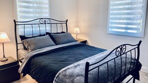 Un letto rifatto con una coperta scura e cuscini, due lampade sui comodini e una finestra con persiane.