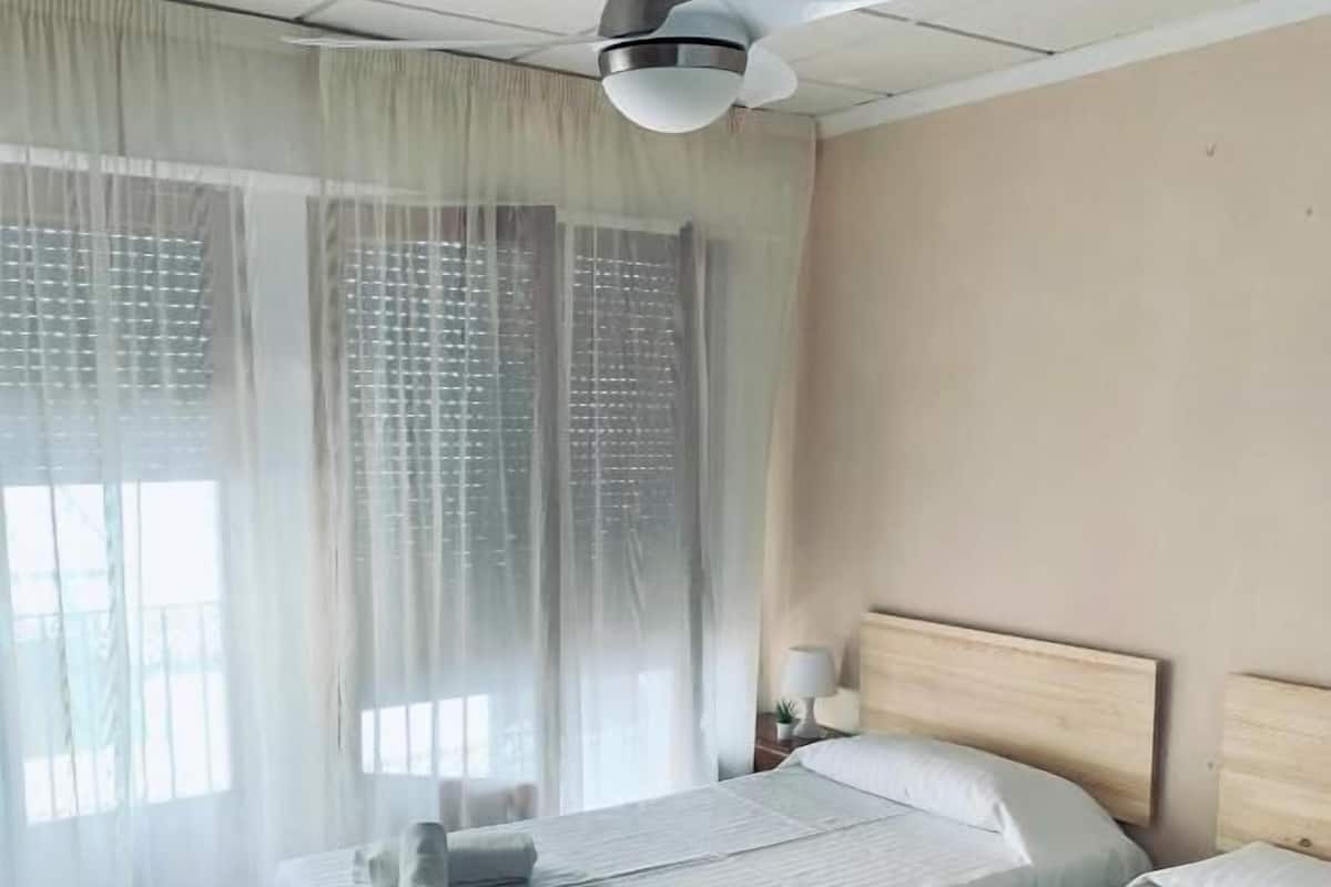 Une chambre d’hôtel avec deux lits, un ventilateur de plafond et une fenêtre avec des rideaux légers.