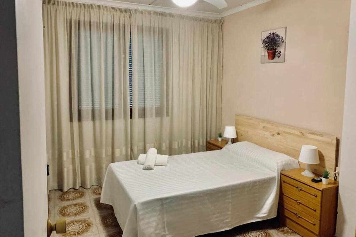 Une chambre d’hôtel avec un lit, deux tables de chevet, un ventilateur de plafond, une fenêtre avec des rideaux et une petite plante en pot accrochée au mur.