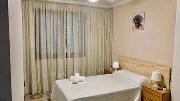 Une chambre d’hôtel avec un lit, deux tables de chevet, un ventilateur de plafond, une fenêtre avec des rideaux et une petite plante en pot accrochée au mur.