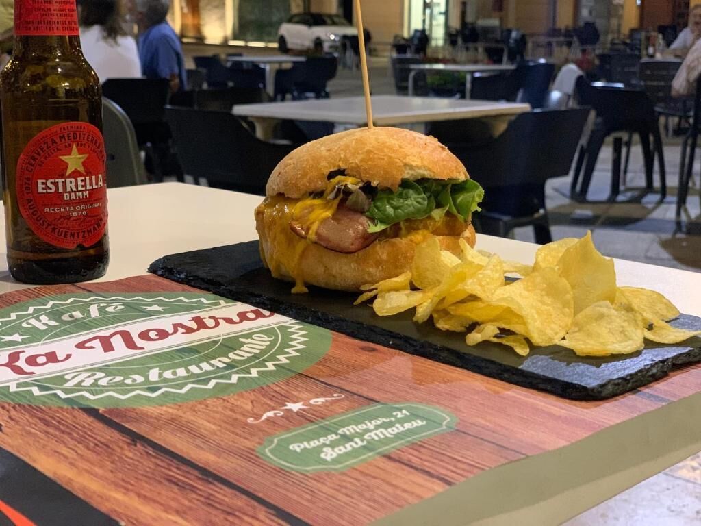 Un sándwich sobre una tabla de madera con patatas fritas, una botella de cerveza Estrella y un letrero de restaurante.