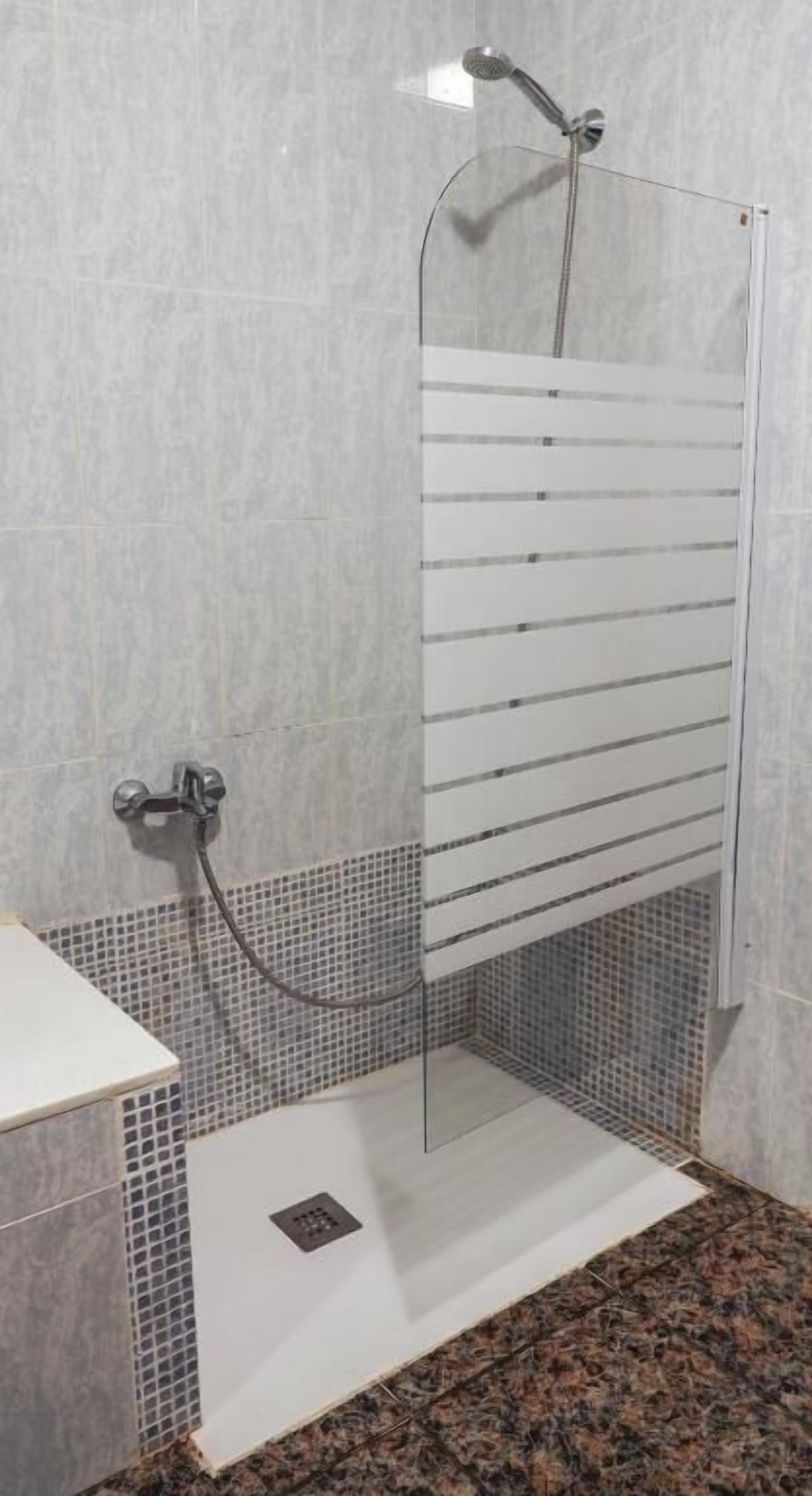 Una ducha con cerramiento de vidrio, suelo de azulejos y grifo empotrado en la pared.