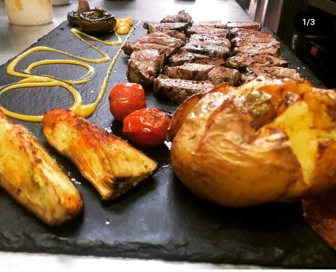 Una tabla de pizarra negra con filete a la parrilla, patatas asadas, tomates y champiñones.