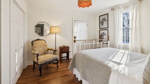 Ein Schlafzimmer mit einem Bett, einem Sessel, einer Lampe, einem Spiegel und einem Fenster mit Vorhängen.
