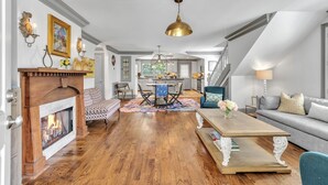 A spacious living room with hardwood floors, a fireplace, a dining area with a table and chairs, and a kitchen in the background.