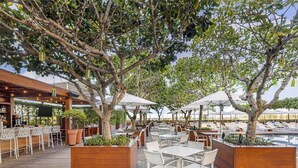 Un área de comedor al aire libre con mesas y sillas de madera, rodeada de árboles y sombrillas.