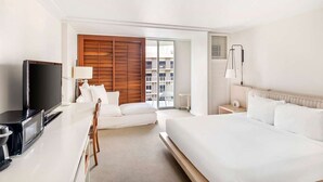 Habitación de hotel con cama, televisor, escritorio con computadora, una silla y vista de la ciudad a través de un ventanal grande.