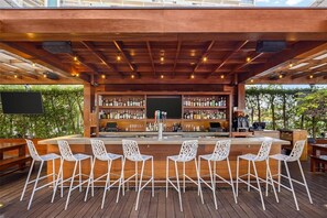 Un área de bar al aire libre con terraza de madera, sillas blancas y una barra llena de botellas.