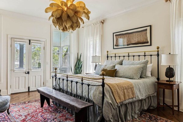 Un dormitorio con cama, banqueta, mesitas de noche, lámpara colgante y ventana con cortinas.