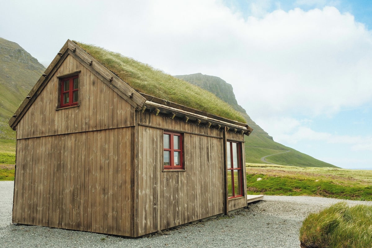 A wooden cabin with a sloped roof and red-framed windows, situated in a grassy landscape with mountains in the background.