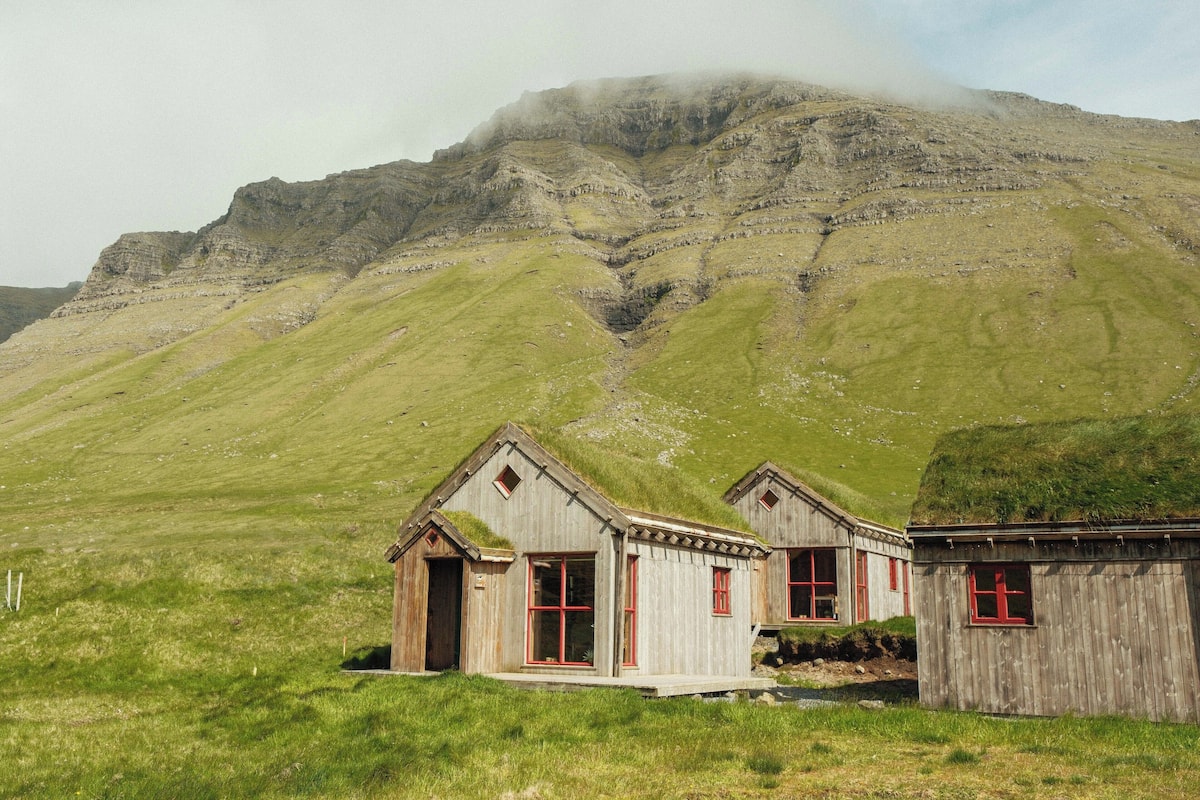 A small, rustic wooden building with a thatched roof, situated on a grassy hillside with a mountain in the background.