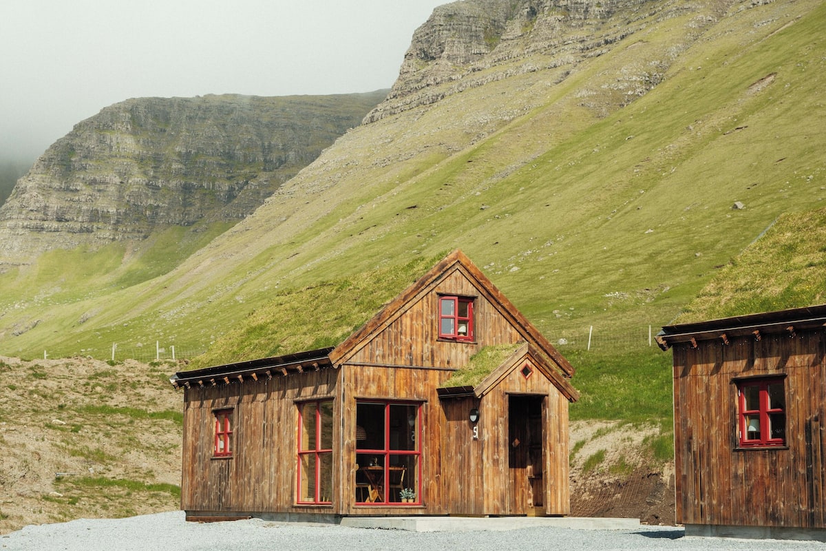 A rustic wooden building with a gabled roof and red-framed windows, situated on a gravel surface with a mountainous backdrop.