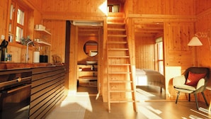 Interior de una cabaña de madera con cocina, baño y un dormitorio con escalera que sube a una buhardilla.