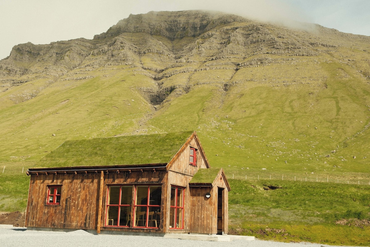 A wooden cabin with a gabled roof and large windows, situated in a grassy valley with a mountain in the background.