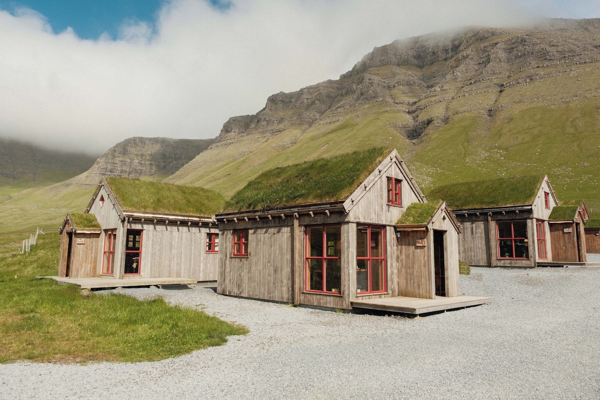 A row of wooden buildings with red-framed windows and a grassy hill in the background.