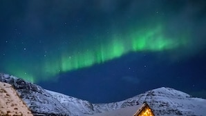 Una cabaña de madera con techo nevado, una ventana con marco rojo y una guirnalda de luces a lo largo de la fachada.