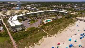 An aerial view of a coastal resort with multiple buildings, a swimming pool, and a beach area with umbrellas.