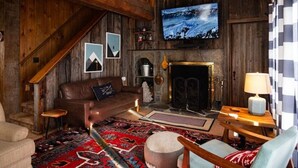 A rustic living room with a wooden staircase, a fireplace, a television mounted on the wall, and a patterned rug.