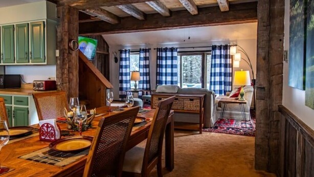 A dining area with a wooden table set for a meal, a staircase leading to a living room with a sofa and a TV, and a kitchen area with cabinets and a microwave.