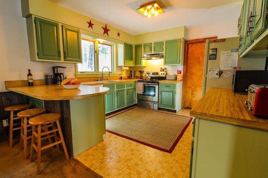 A kitchen with green cabinets, a wooden countertop, a sink, a stove, and a refrigerator. There are three stools near the sink.