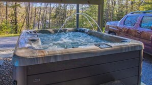 A hot tub with a built-in seat and a water spout, surrounded by a covered area with a view of a forested area and a parked vehicle.