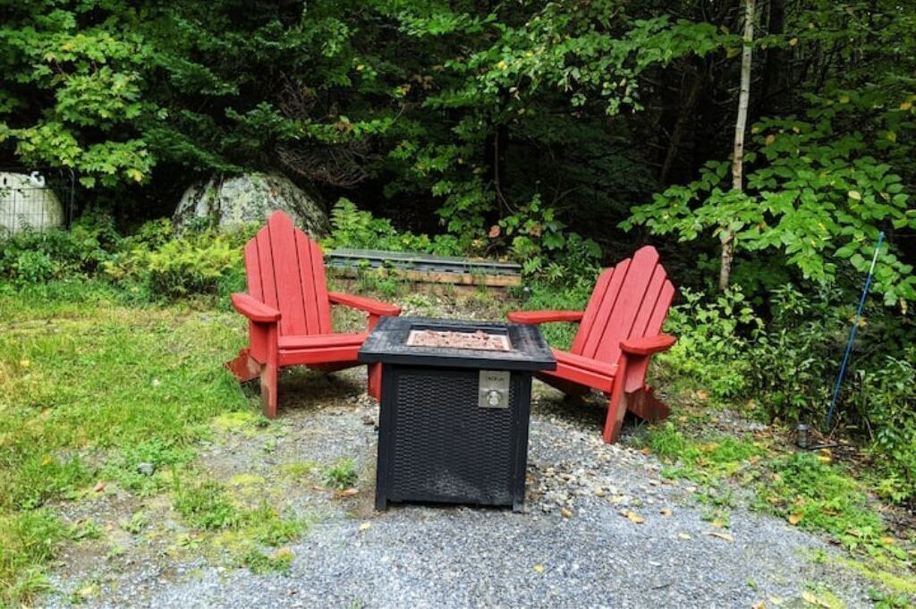 Un sillón Adirondack rojo dispuesto alrededor de una pequeña mesa negra con una placa metálica en la parte superior, ubicado en un sendero de grava rodeado de follaje.