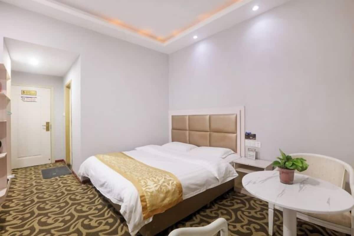 Une chambre d’hôtel avec un lit, une table de chevet agrémentée d’une plante, une table ronde et une moquette à motifs.