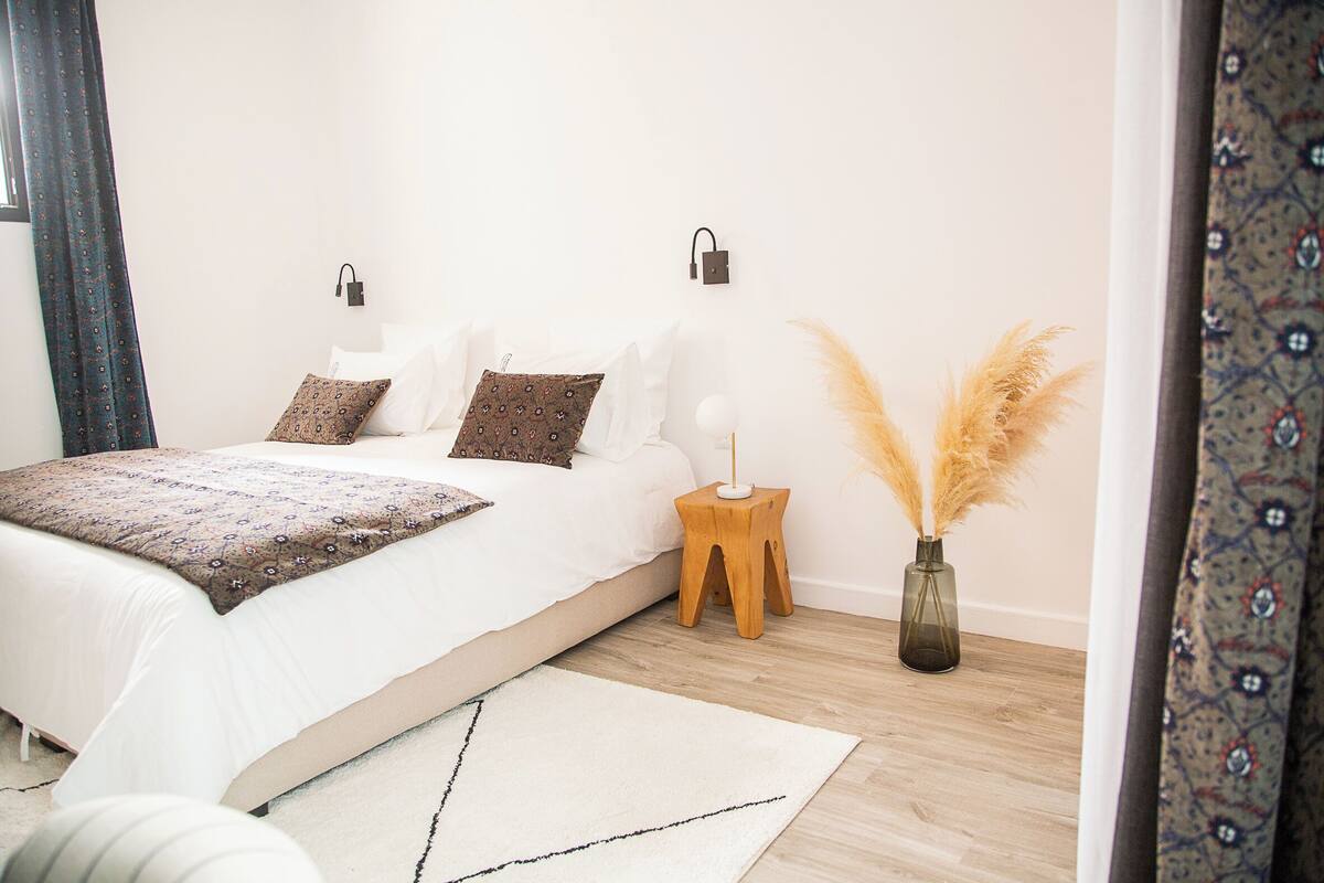 Un dormitorio con una cama, lámparas de noche, una mesita de noche, un jarrón con plumas de pampa y una alfombra estampada.