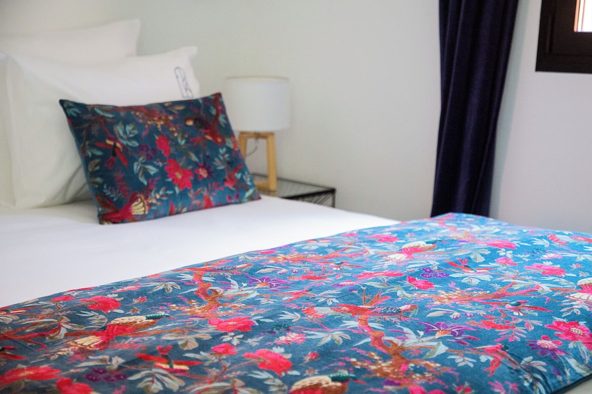 Una habitación de hotel con una cama, una colcha estampada con motivos florales, un almohadón blanco, una lámpara de noche y una cortina oscura.