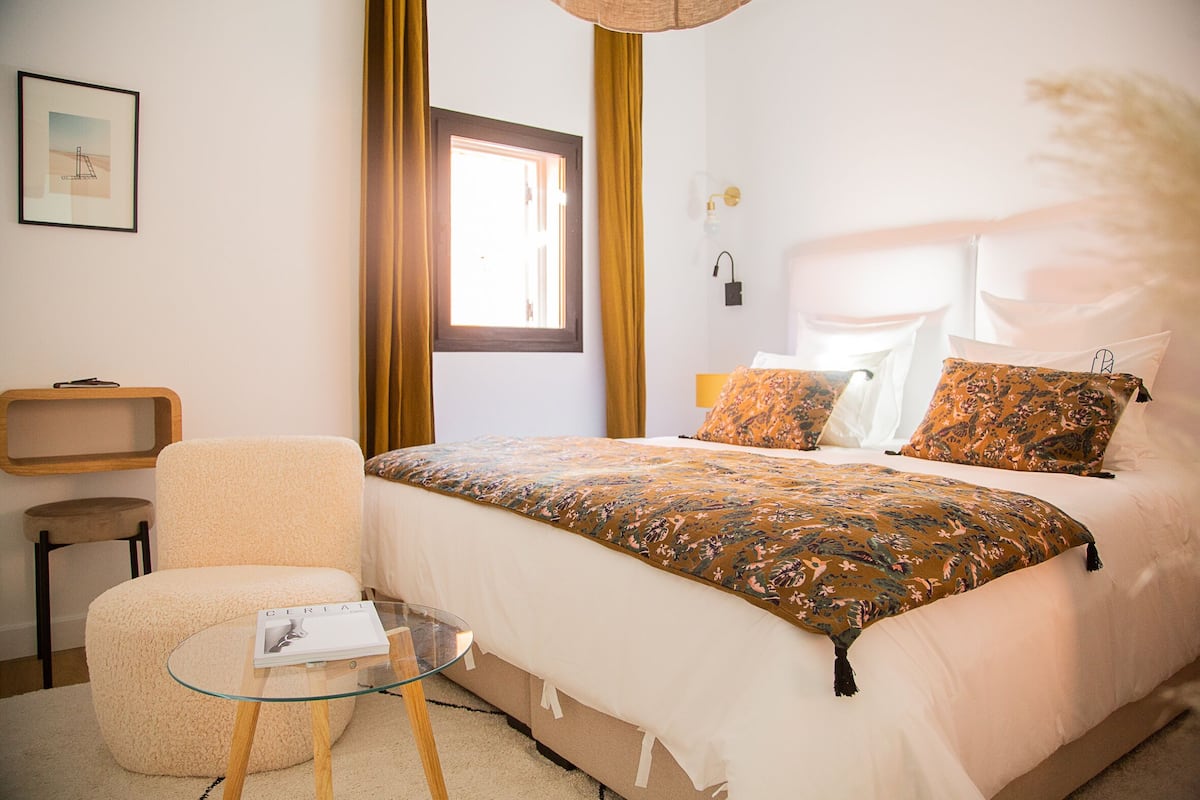 Un dormitorio ordenado con una cama, una silla, una mesita de cristal y una ventana con cortinas.