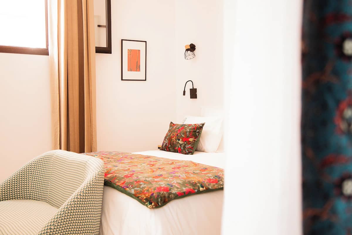 Un dormitorio con cama, una silla, una lámpara de pared y un cuadro enmarco.