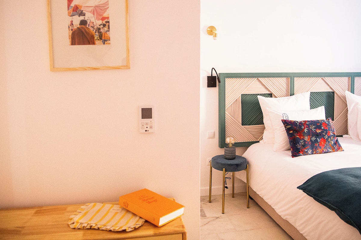 Un dormitorio ordenado con cama, mesita de noche y un taburete pequeño.