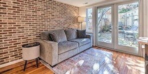Ein Zimmer mit einer Backsteinwand, einem grauen Sofa, einer Lampe und Blick durch eine Glastür auf einen Außenbereich.