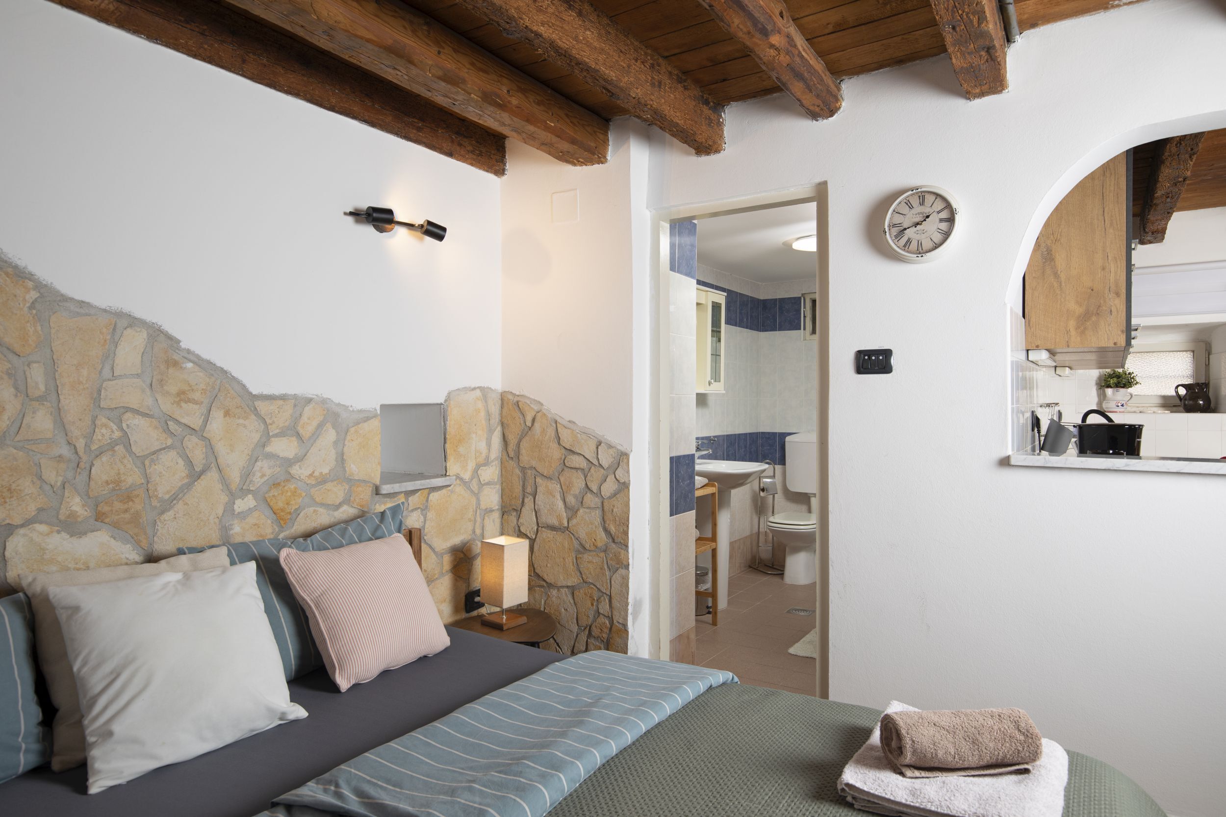 Un dormitorio con cabecera de piedra, cama con ropa de cama a rayas, lámpara de noche y una toalla sobre la cama.