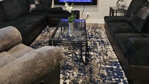 Un salon avec un téléviseur à écran plat fixé au mur, deux canapés noirs, une table basse en verre ornée d’un vase et un tapis à motifs.