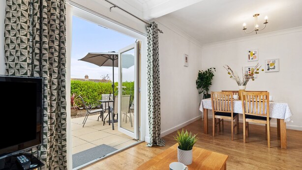 A living room with a TV, wooden furniture, a dining table with chairs, and a view of a garden through a sliding door. - St Helens Apartment 2 - Short Walk to Beach (Wisemans Bridge)