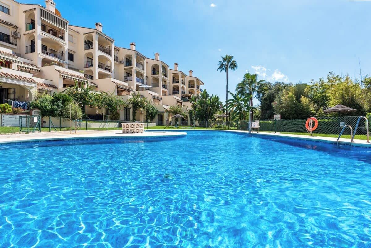 A swimming pool surrounded by a multi-story building with balconies, palm trees, and a clear blue sky.