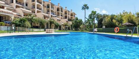 A swimming pool surrounded by a multi-story building with balconies, palm trees, and a clear blue sky.