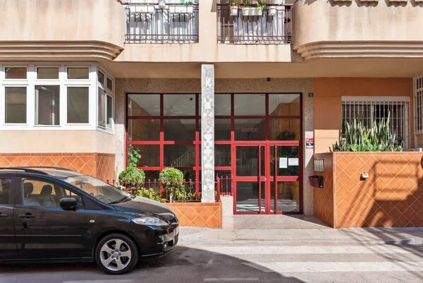 Un bâtiment avec une porte d’entrée rouge, une voiture garée devant et un petit jardin avec des plantes.