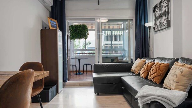 A modern living room with a sofa, a dining table, and chairs. There is a large window with curtains, a potted plant, and a clock on the wall.