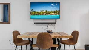 A dining area with a wooden table, two brown chairs, and a television displaying a tropical beach scene.