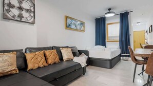 A modern living room with a black sofa, a bed, a dining area, and a large wall clock.