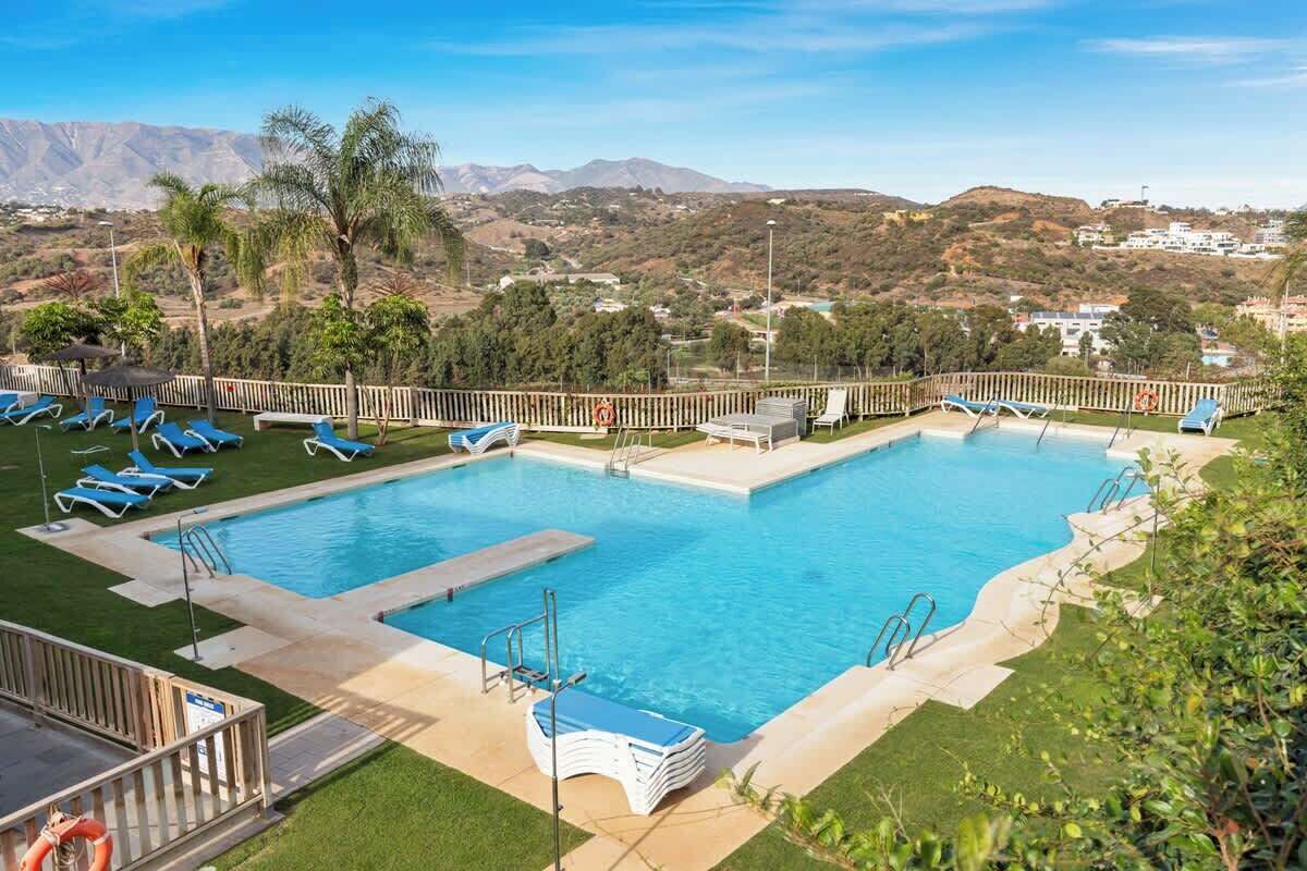 A swimming pool with blue lounge chairs, a lifeguard stand, and a view of hills and buildings in the background.
